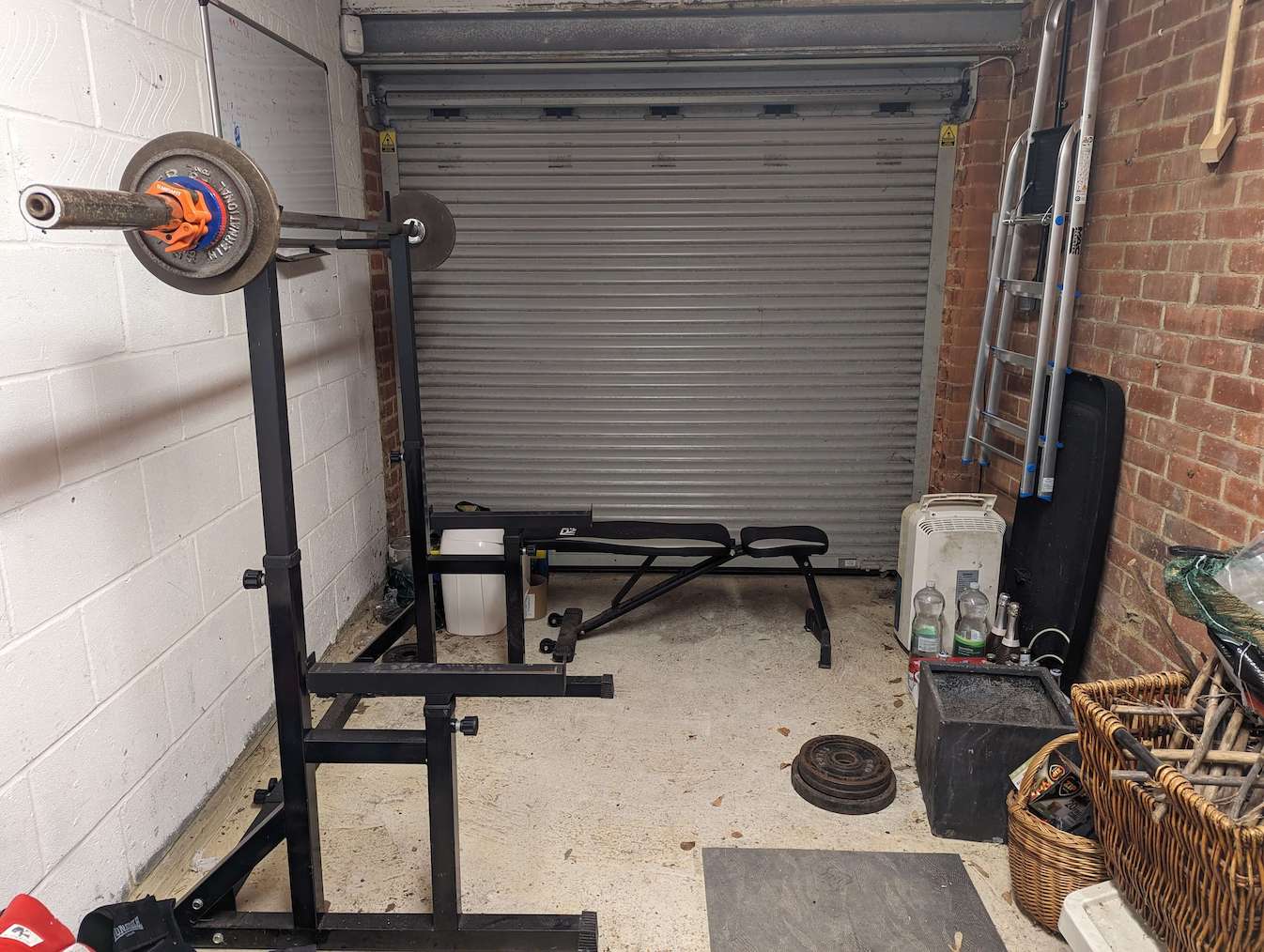 Messy home garage gym showing a squat rack holding up a barbell weight plates on either end; a bench in the background; and additional plates on the floor next to the rack